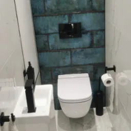 Nowoczesna łazienka z turkusowymi, cegiełkowymi płytkami na ścianie, wiszącą toaletą, białą umywalką z czarną armaturą i marmurową podłogą.