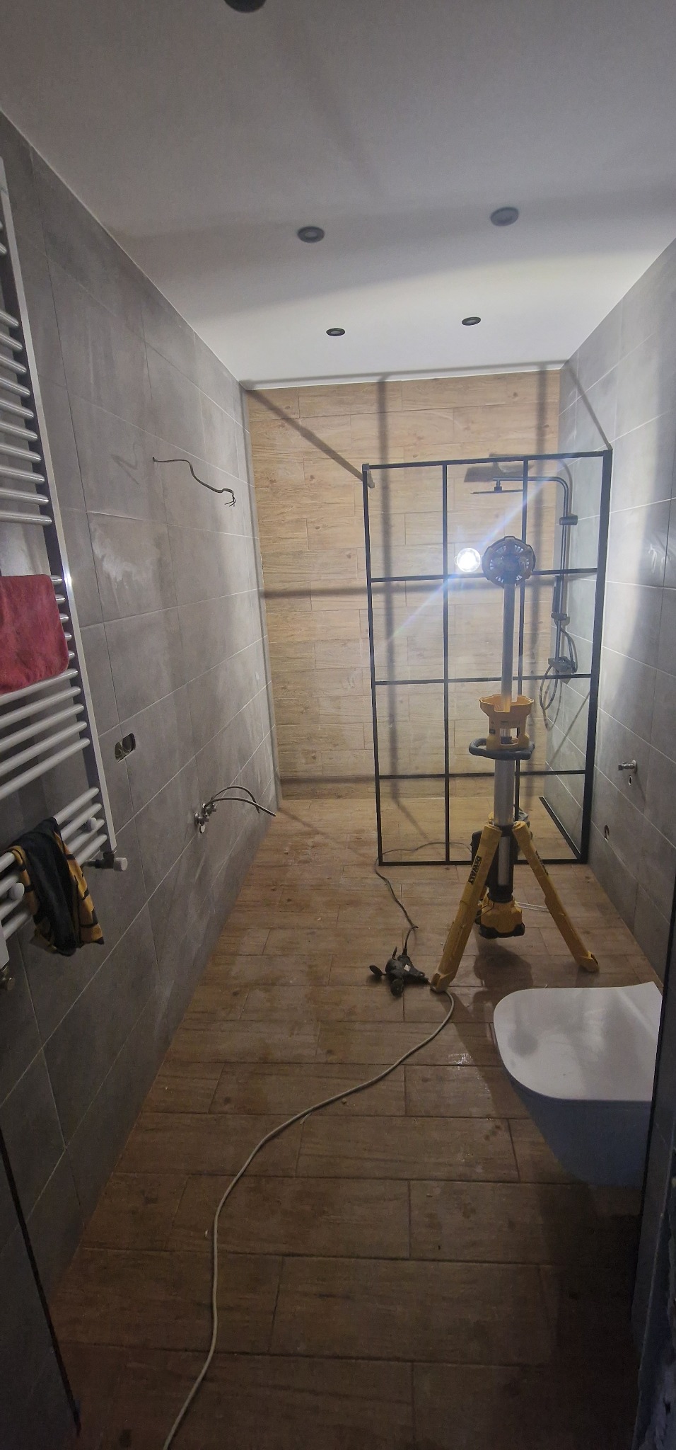 Łazienka w trakcie remontu: zamontowana kabina prysznicowa z czarnymi profilami, widoczna lampa budowlana na statywie oświetlająca wnętrze, płytki imitujące drewno na podłodze, częściowo położone...