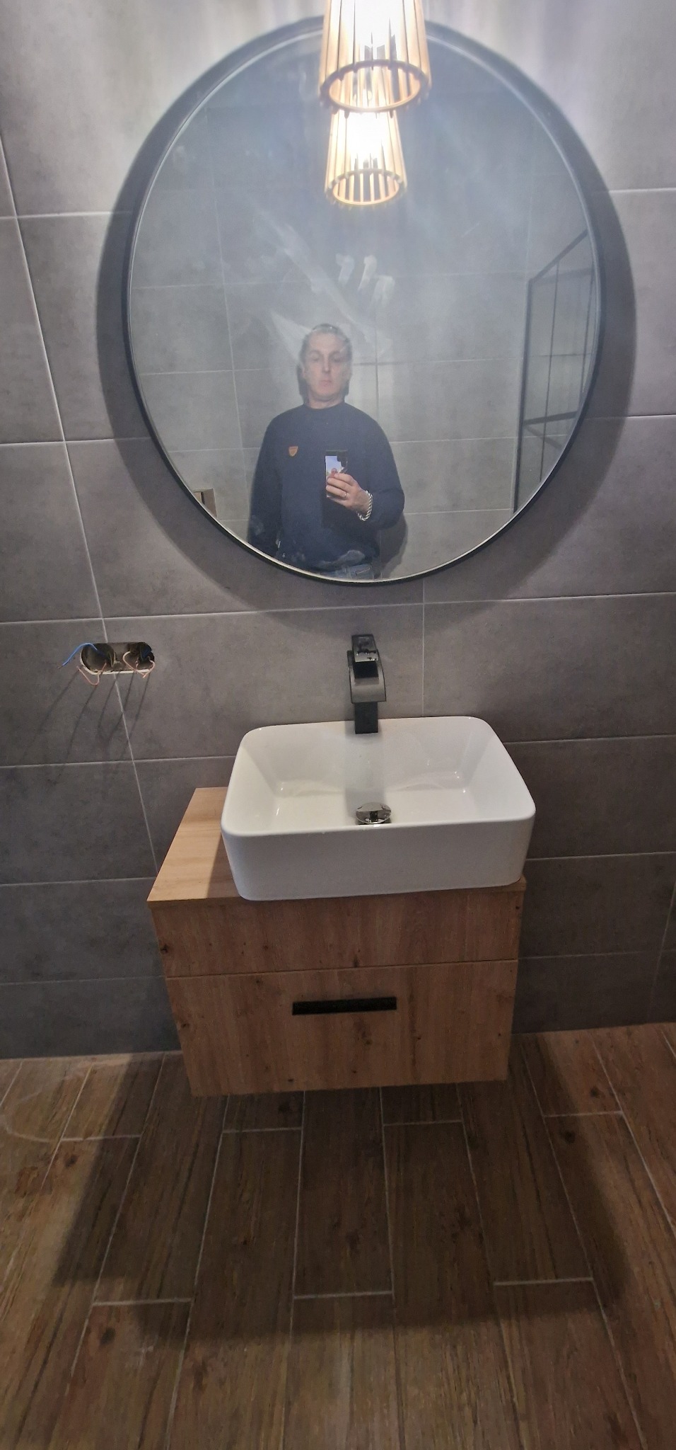 Nowoczesna łazienka z umywalką nablatową na drewnianej szafce, ciemnoszara glazura ścienna, okrągłe lustro z czarną ramą i lampa wisząca, widoczne niedokończone podłączenie elektryczne do oświetlenia.