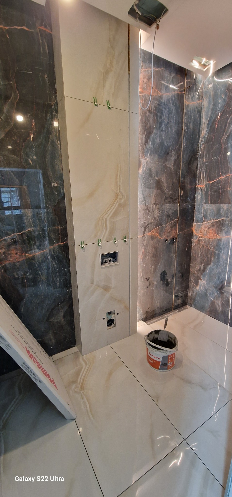 Łazienka w trakcie wykańczania: marmurowe płytki na ścianach i podłodze, widoczna puszka podtynkową WC, poziomowanie płytek za pomocą klipsów, wiadro z zaprawą klejową.