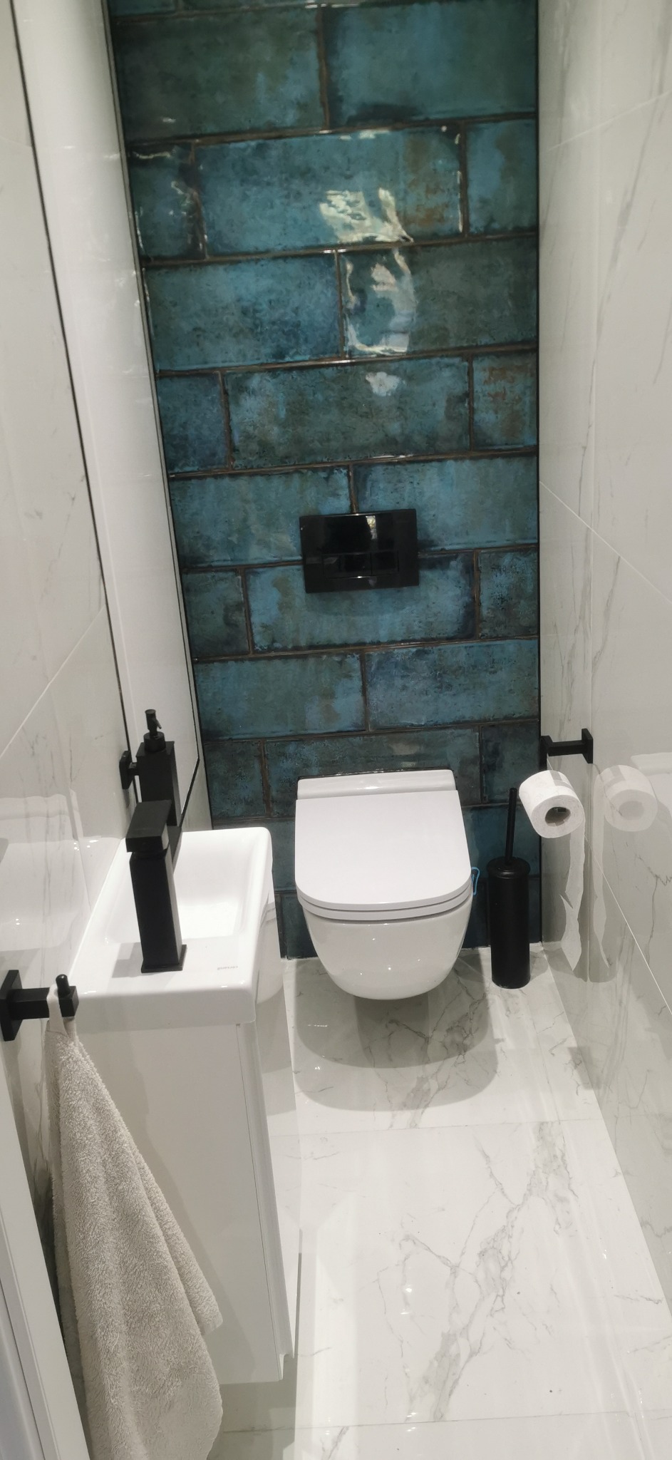 Nowoczesna łazienka z turkusowymi, cegiełkowymi płytkami na ścianie, wiszącą toaletą, białą umywalką z czarną armaturą i marmurową podłogą.