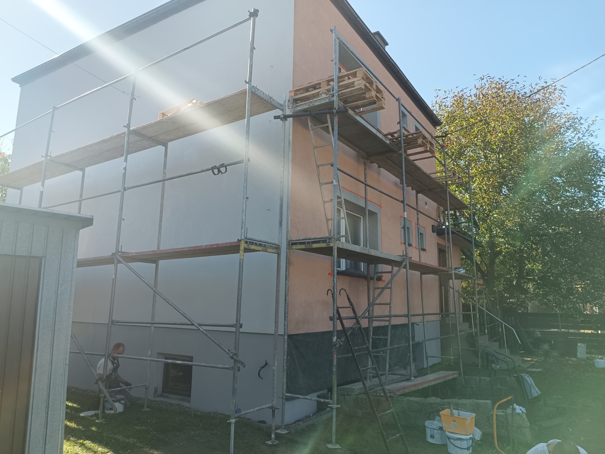 Dwukondygnacyjny dom w trakcie prac elewacyjnych: część ściany pokryta szarą warstwą, druga część w odcieniach pomarańczy, rusztowania z paletami, pracownik przy gruncie z wiadrem farby, słoneczny...