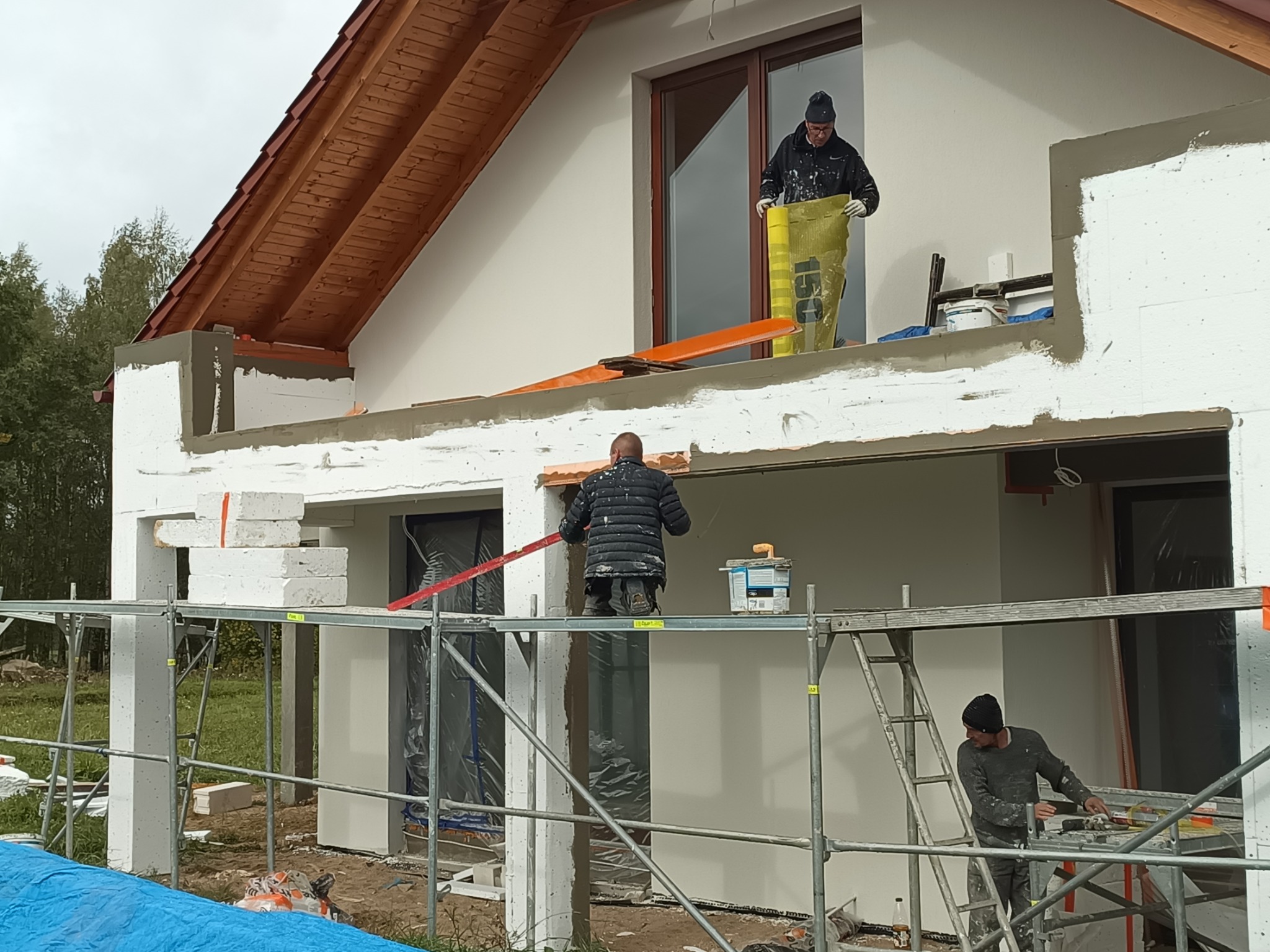 Prace wykończeniowe na elewacji domu jednorodzinnego, widoczne rusztowania, pracownicy w trakcie nakładania zaprawy i przygotowywania materiałów, ściany z białego bloczka częściowo pokryte szarą...