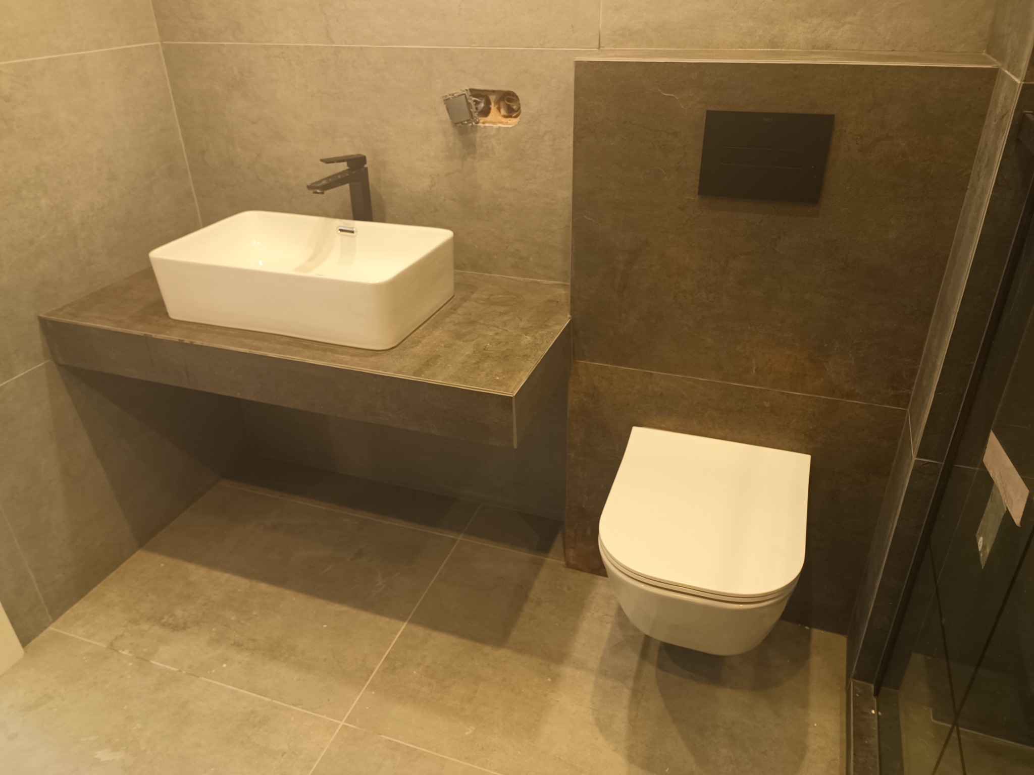 Nowoczesna łazienka z umywalką nablatową na szarym blacie, wiszącą toaletą i szarą armaturą, widoczne niedokończone podłączenie elektryczne.