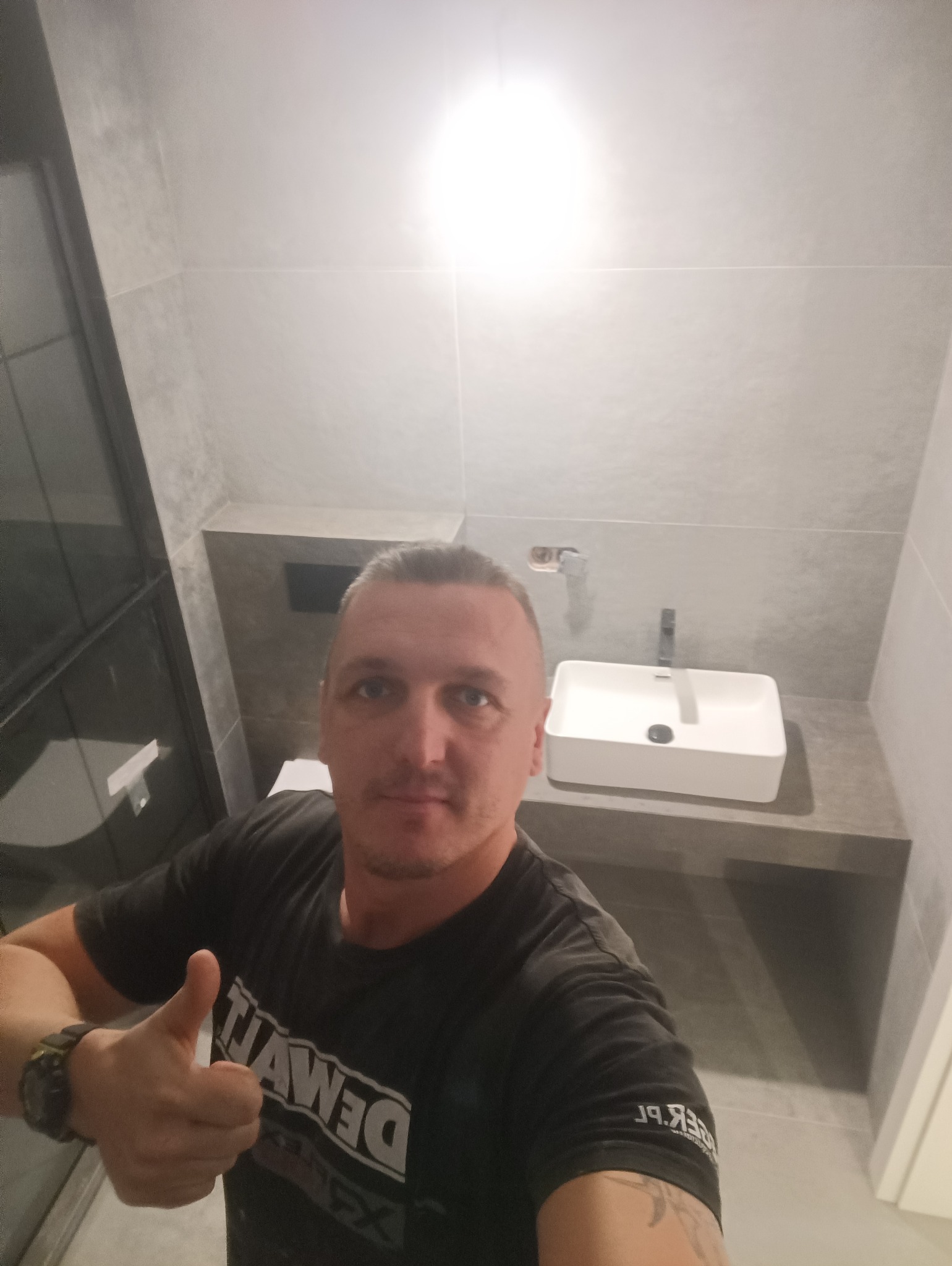 Uśmiechnięty mężczyzna w koszulce Dewalt, z kciukiem w górę, na tle wykończonej łazienki z szarymi płytkami i białą umywalką.