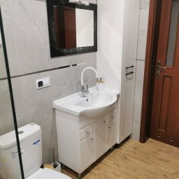 Usługi remontowo-budowlane - Nowoczesna łazienka z umywalką na szafce z chromowanymi uchwytami, toaletą z widoczną naklejką informacyjną, lustrem w czarnej, zdobionej ramie i wysoką białą szafką na tle szarych płytek...