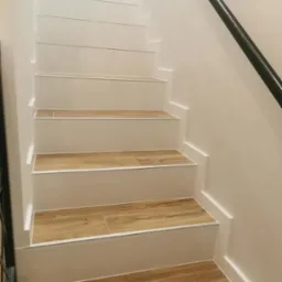 Schody wykończone płytkami imitującymi drewno, białe stopnice, czarna metalowa poręcz, minimalistyczny design.