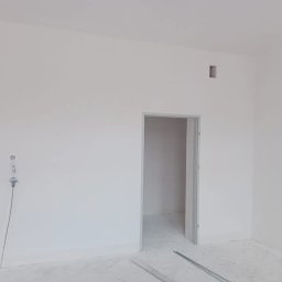 Usługi remontowo-budowlane - Wnętrze pokoju po malowaniu w Leżajsku, widoczne świeżo pomalowane białe ściany, okno, puste gniazdko elektryczne oraz metalowe listwy na podłodze.