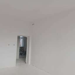 Usługi remontowo-budowlane - Świeżo pomalowane na biało wnętrze z widocznymi puszkami elektrycznymi, w tle otwarte drzwi do kolejnego pomieszczenia z widocznymi narzędziami i drabiną.