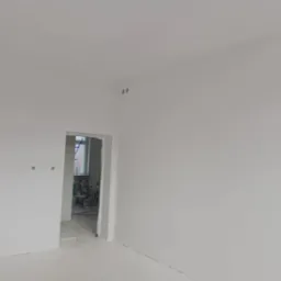 Świeżo pomalowane na biało wnętrze z widocznymi puszkami elektrycznymi, w tle otwarte drzwi do kolejnego pomieszczenia z widocznymi narzędziami i drabiną.