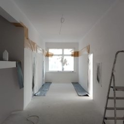 Usługi remontowo-budowlane - Korytarz w trakcie przygotowań do malowania: ściany zabezpieczone folią i taśmą malarską, podłoga osłonięta, drabina aluminiowa po prawej.