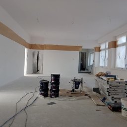 Usługi remontowo-budowlane - Przygotowane do malowania wnętrze z zaklejonymi taśmą odcięciami i zabezpieczonymi folią oknami, widoczne wiaderka z farbą Dulux, agregat malarski i opakowania Viessmann.