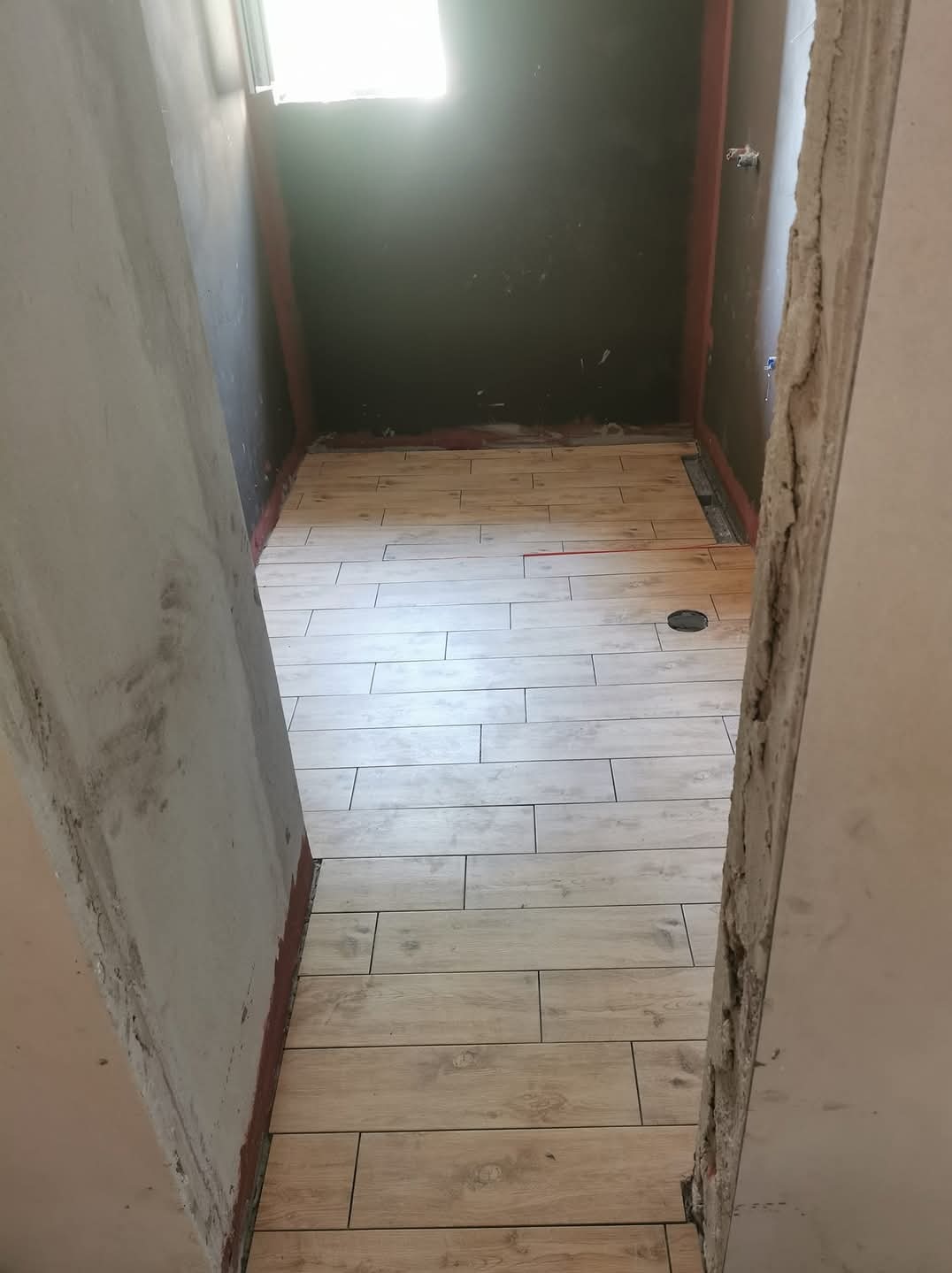 Układanie podłogowych płytek imitujących drewno w małej łazience z odpływem liniowym, widok z korytarza, ściany w trakcie przygotowania.