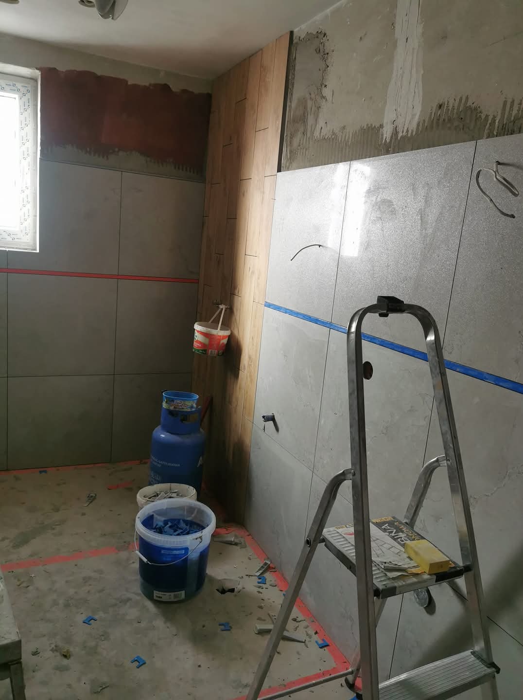 Łazienka w trakcie remontu: szare płytki imitujące kamień na ścianach, drewnopodobne panele, niebieska taśma malarska wyznaczająca linie, drabina z żółtą gąbką, butla gazowa i wiadro z niebieskimi...