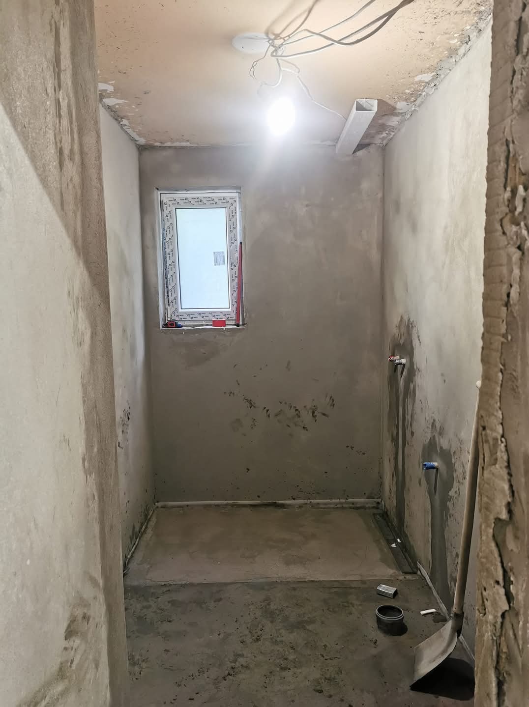 Surowa łazienka w trakcie prac wykończeniowych: ściany pokryte szarą zaprawą, nowe okno PCV z poziomnicą, wystające przewody elektryczne, przygotowane instalacje wodne, widoczna łopata i puszka...
