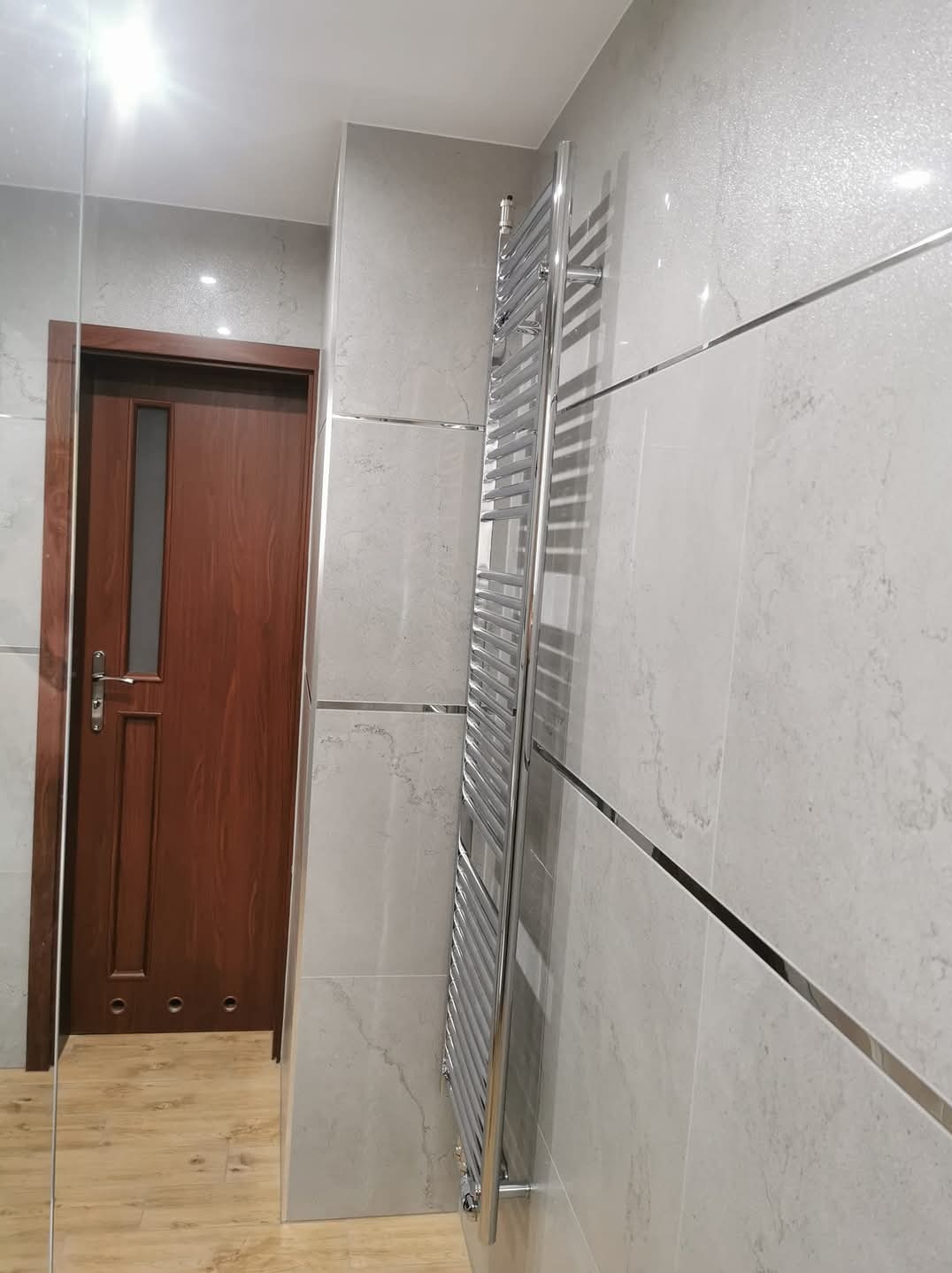 Wykończona łazienka z szarymi płytkami imitującymi marmur, chromowanym grzejnikiem drabinkowym i drewnianymi drzwiami z przeszklonym panelem.