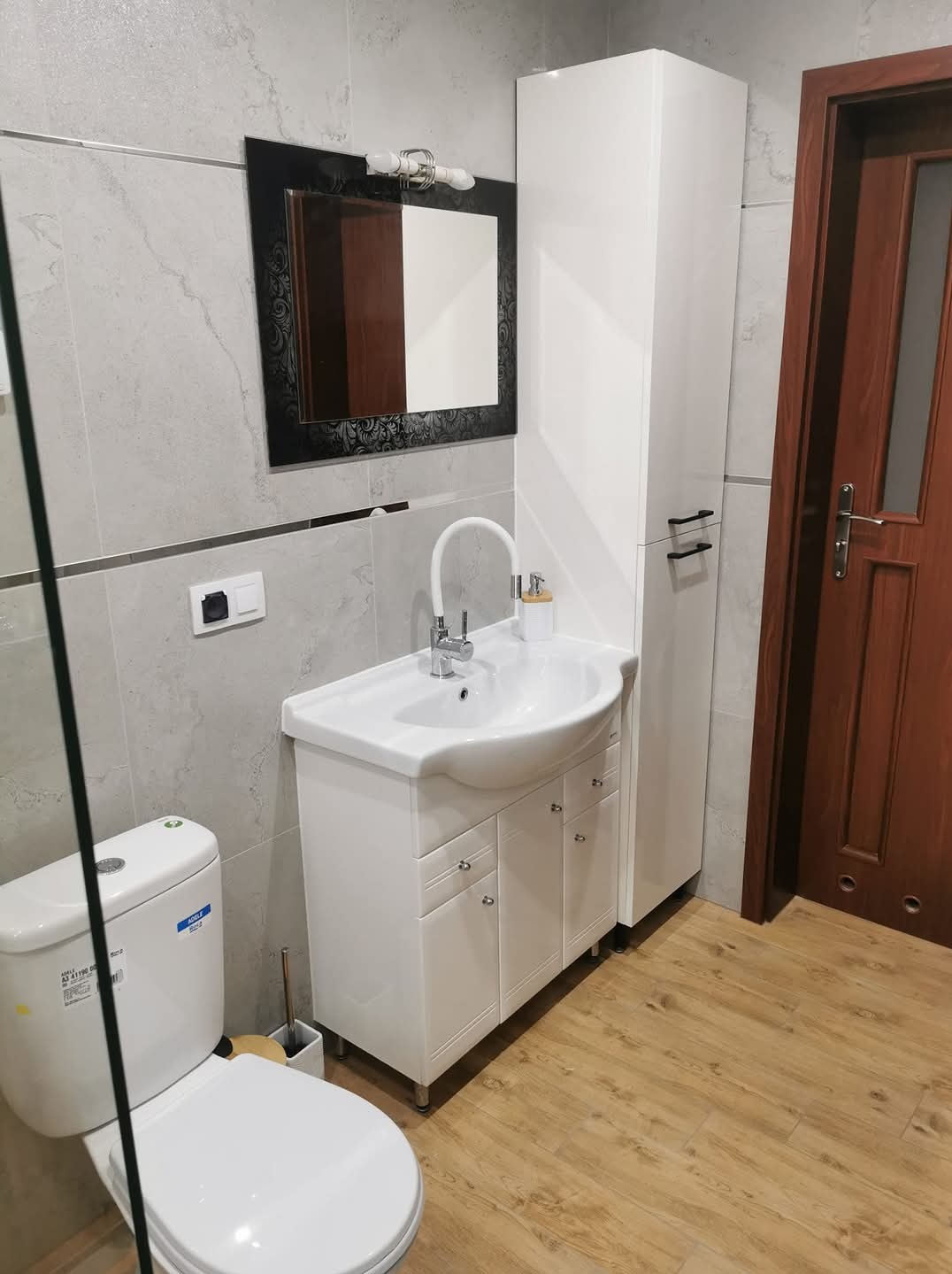 Nowoczesna łazienka z umywalką na szafce z chromowanymi uchwytami, toaletą z widoczną naklejką informacyjną, lustrem w czarnej, zdobionej ramie i wysoką białą szafką na tle szarych płytek...