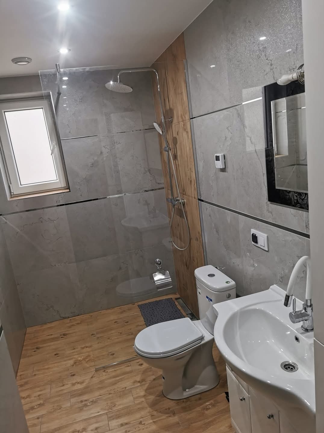 Nowoczesna łazienka z szarymi płytkami imitującymi kamień, drewnopodobną wstawką przy kabinie prysznicowej, drewnianą podłogą, białą ceramiką i czarnym lustrem z ozdobnymi detalami.