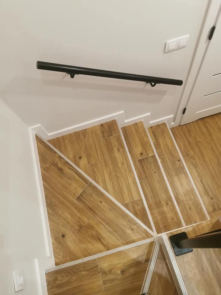 Klatka schodowa wykończona panelami imitującymi drewno, z czarną, minimalistyczną poręczą zamontowaną na białej ścianie. Widoczny fragment białych drzwi z delikatnym frezowaniem oraz włącznik światła.