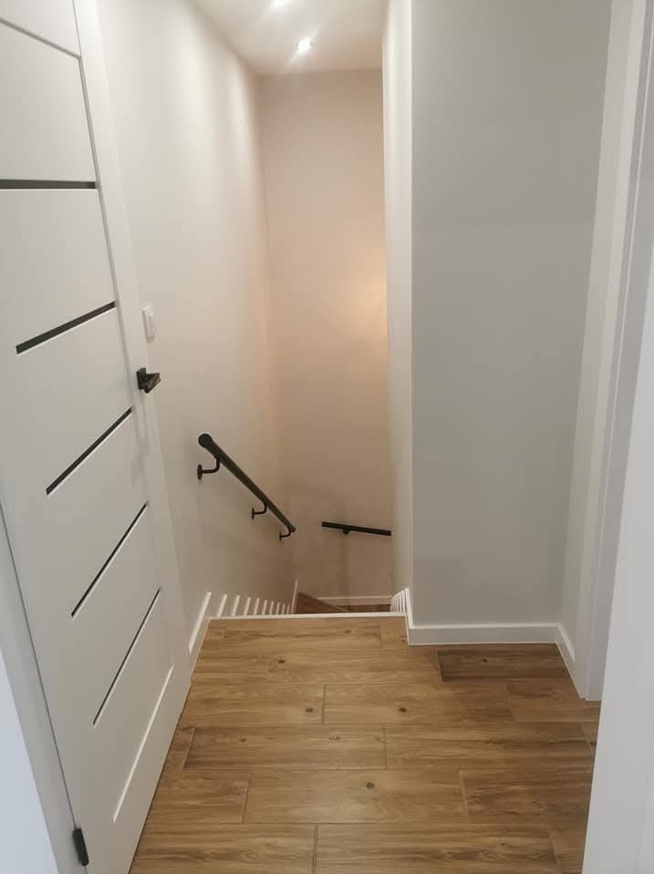 Korytarz z drewnopodobną podłogą, białe ściany i schody w dół z czarną poręczą, widoczna biała, nowoczesna drzwi z czarnymi poziomymi akcentami.