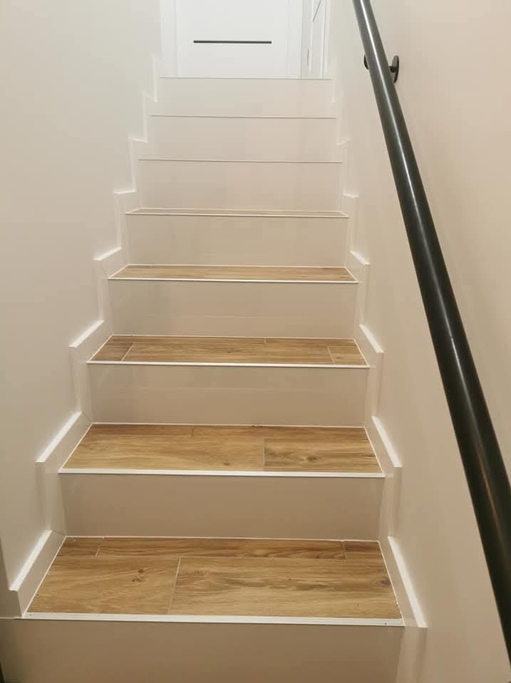 Schody wewnętrzne z drewnopodobnymi płytkami na stopniach, białe ściany i czarna poręcz po prawej stronie.