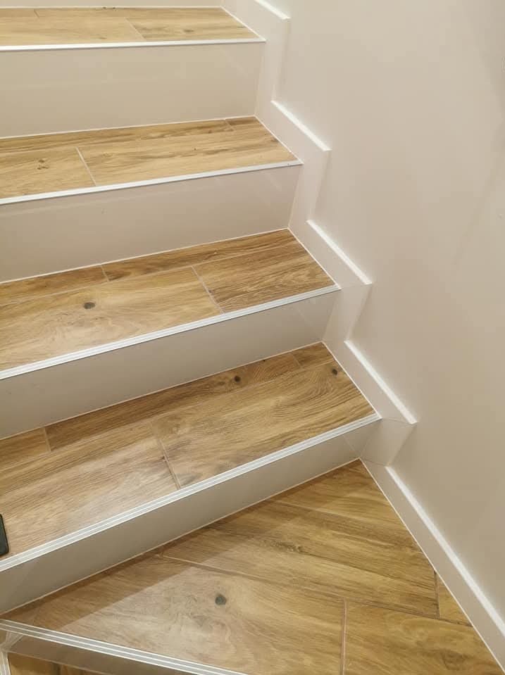 Wykończone schody z drewnopodobnymi płytkami, białe stopnice i listwy przypodłogowe, widoczne łączenia płytek.