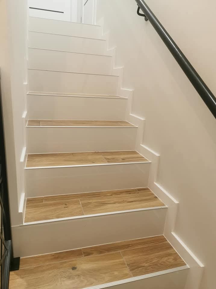 Schody wykończone płytkami imitującymi drewno, białe stopnice, czarna metalowa poręcz, minimalistyczny design.