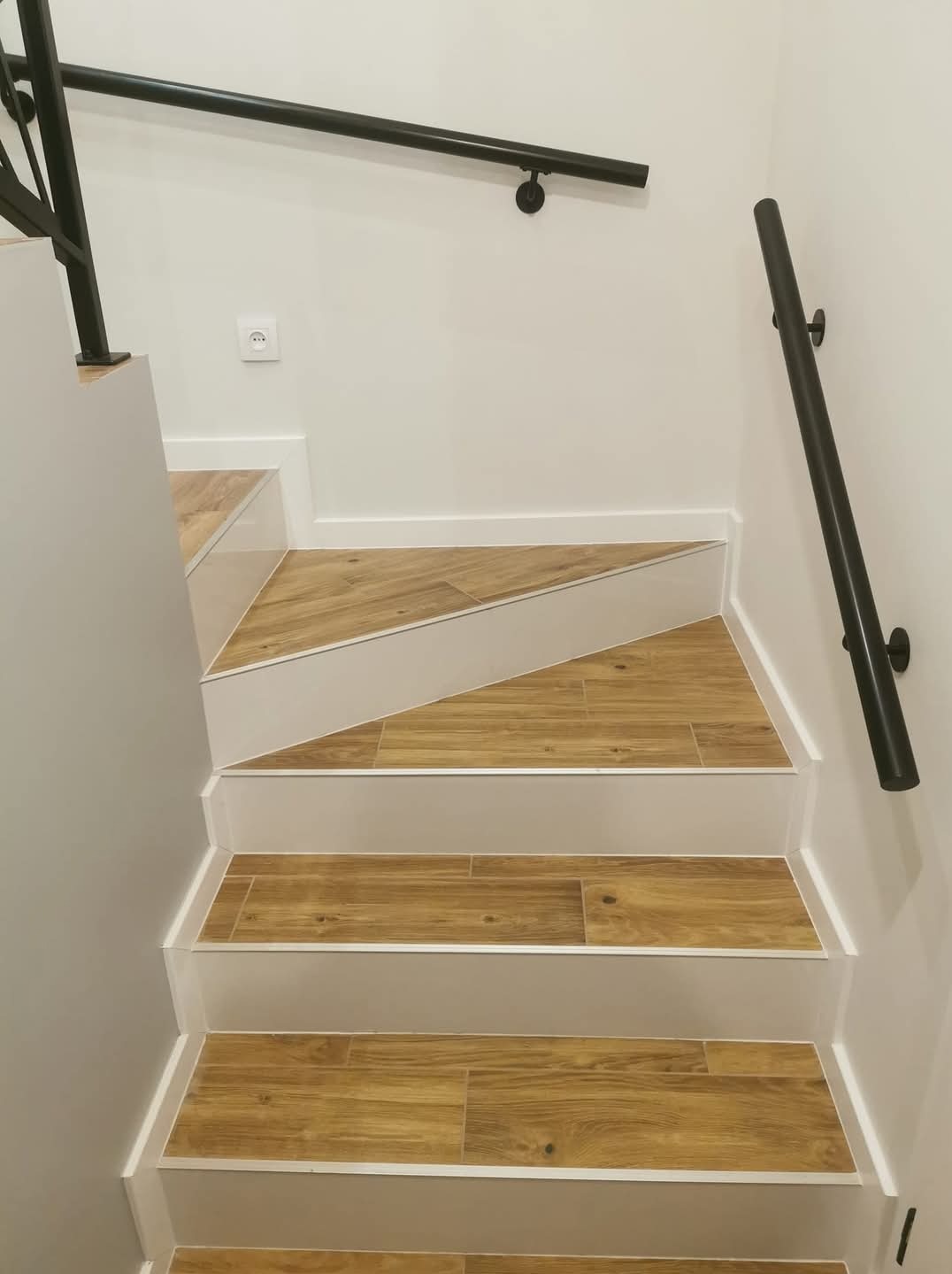 Schody wyłożone płytkami imitującymi drewno, z czarną metalową balustradą przy białej ścianie, widoczne z góry, z detalem w postaci gniazdka elektrycznego na ścianie.