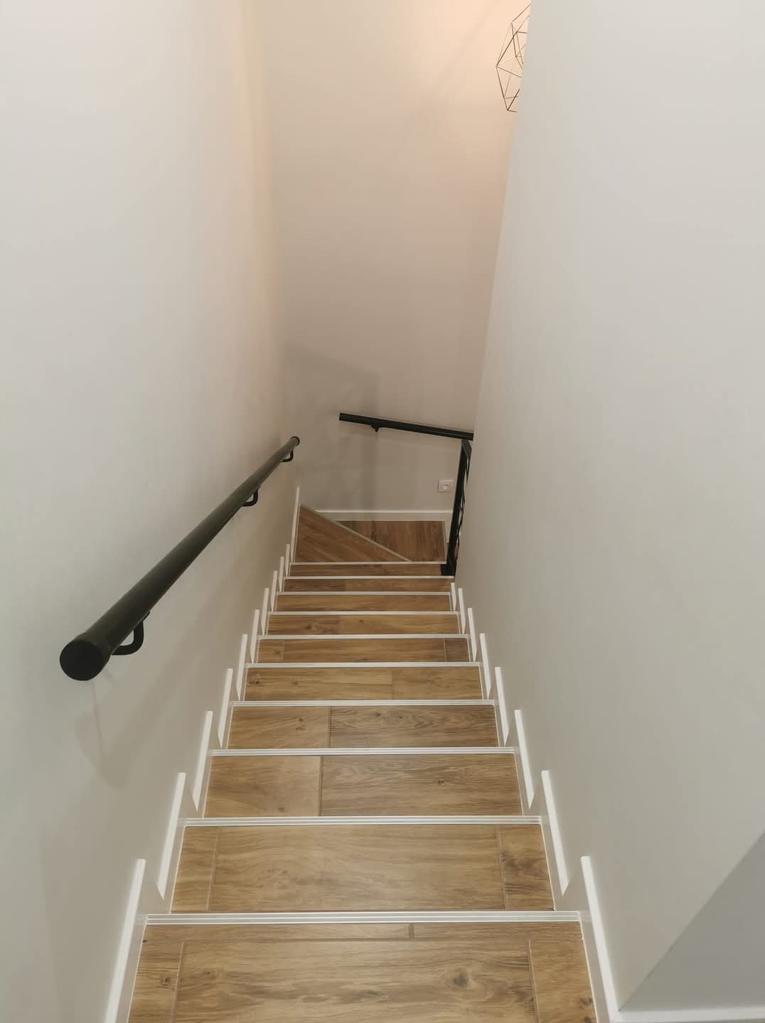 Schody wyłożone płytkami imitującymi drewno, z czarną poręczą po lewej stronie, widok z góry na dół, białe ściany.