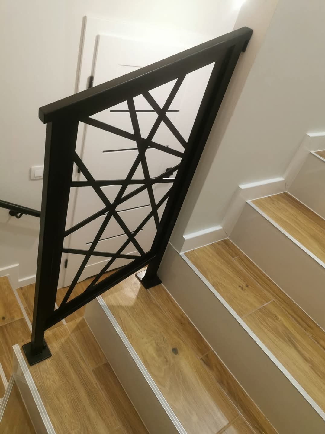 Nowoczesna balustrada schodowa z czarnego metalu o geometrycznym wzorze na tle drewnopodobnych płytek schodowych i białej ściany z drzwiami.