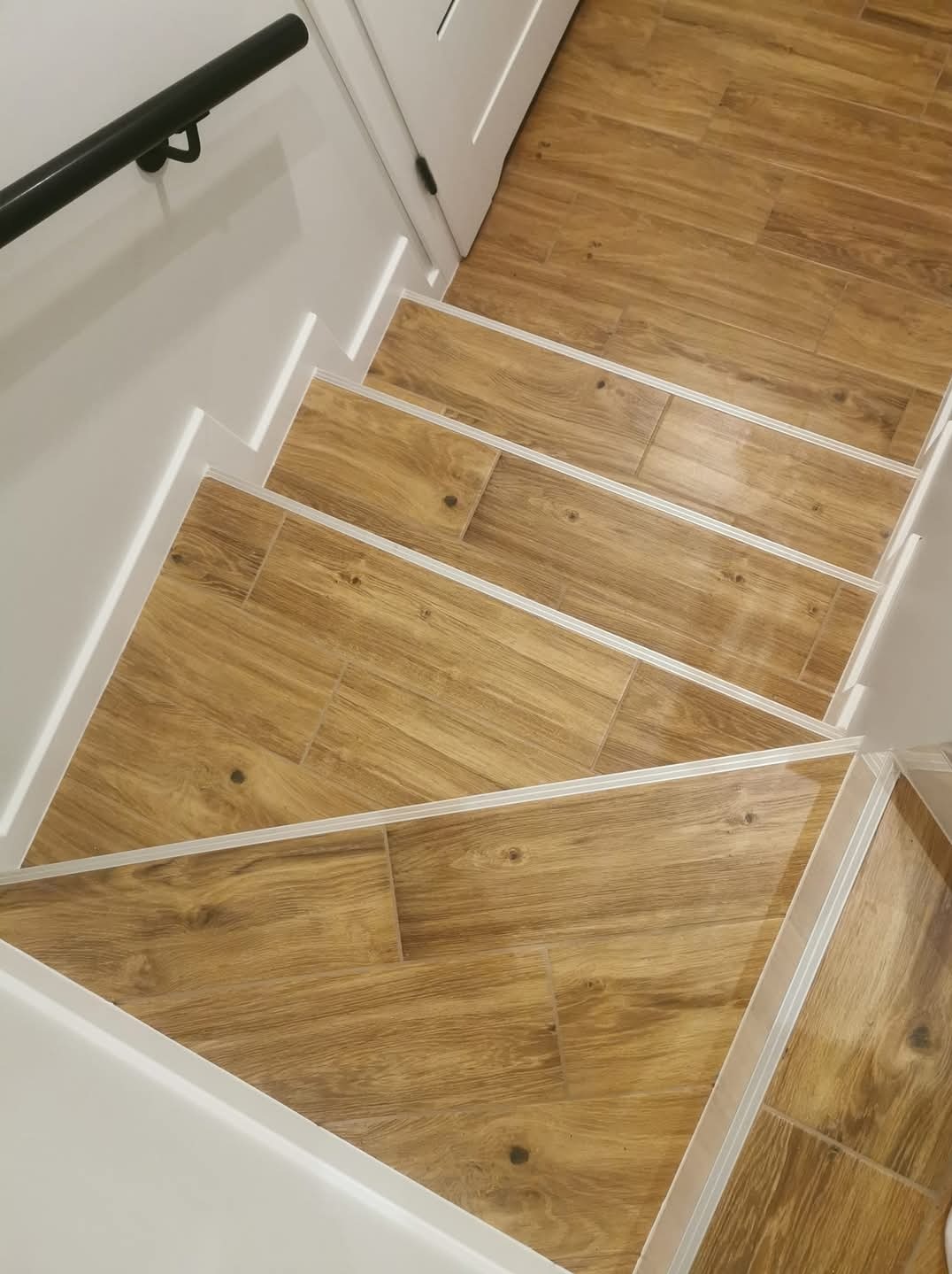 Schody wyłożone płytkami imitującymi drewno, z białymi listwami przypodłogowymi i czarną poręczą, prowadzące do białych drzwi.