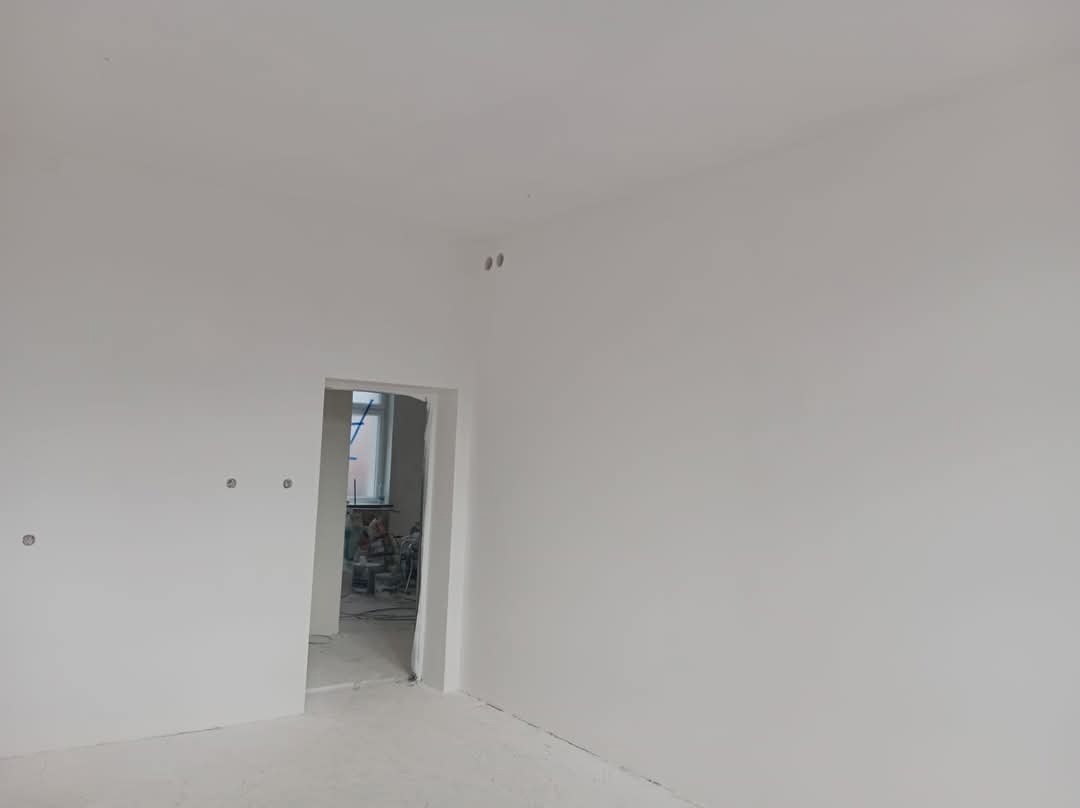 Świeżo pomalowane na biało wnętrze z widocznymi puszkami elektrycznymi, w tle otwarte drzwi do kolejnego pomieszczenia z widocznymi narzędziami i drabiną.