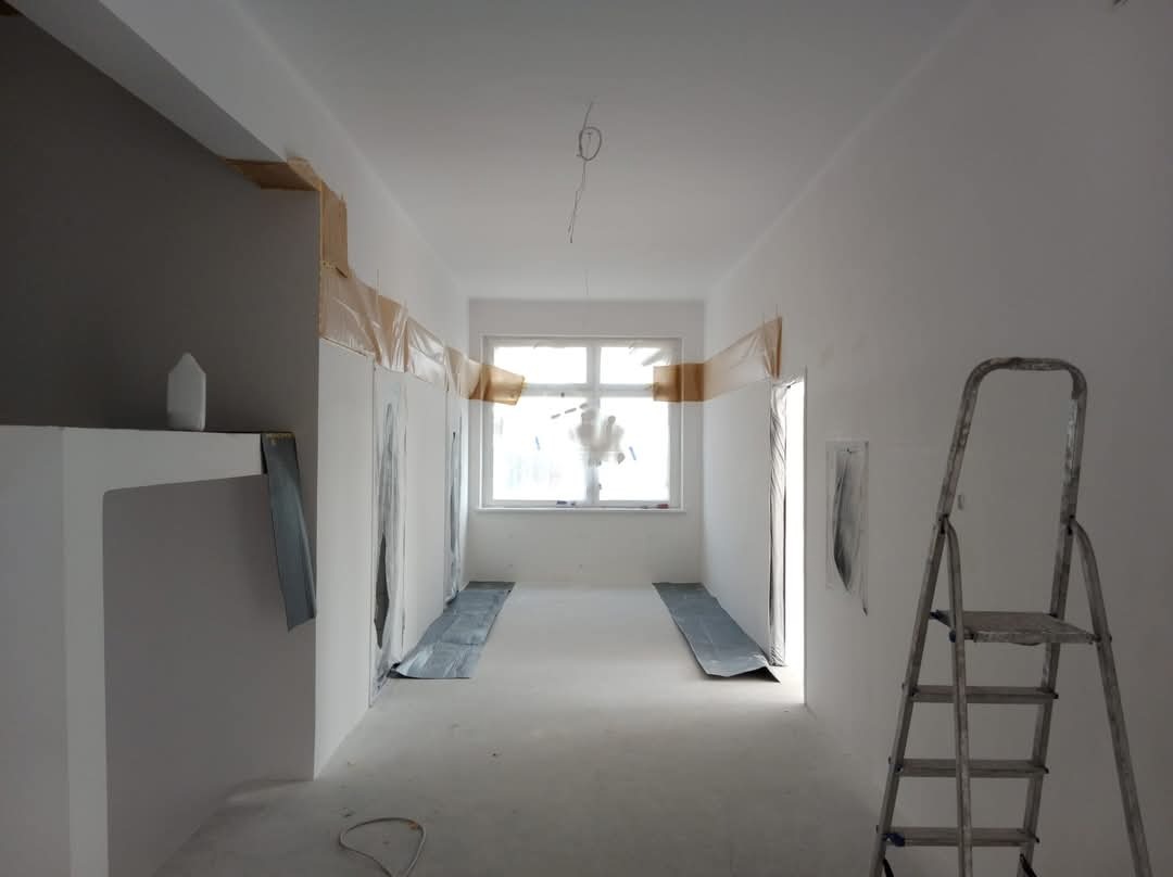 Korytarz w trakcie przygotowań do malowania: ściany zabezpieczone folią i taśmą malarską, podłoga osłonięta, drabina aluminiowa po prawej.