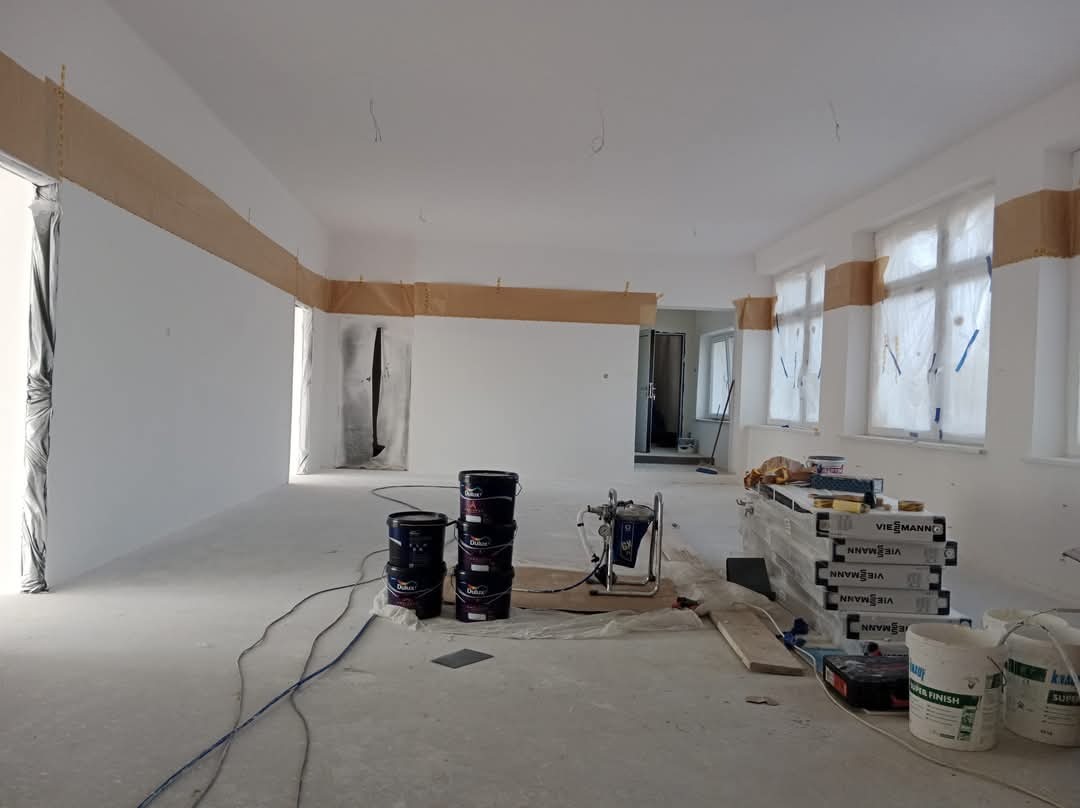 Przygotowane do malowania wnętrze z zaklejonymi taśmą odcięciami i zabezpieczonymi folią oknami, widoczne wiaderka z farbą Dulux, agregat malarski i opakowania Viessmann.