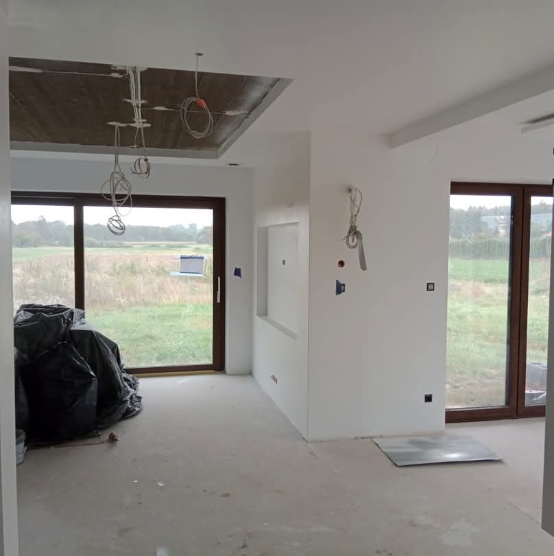 Wnętrze w trakcie remontu: gładkie, białe ściany, sufit z odsłoniętą konstrukcją i zwisającymi kablami, duże okna z widokiem na łąkę, podłoga przygotowana do wykończenia, zabezpieczona folią...