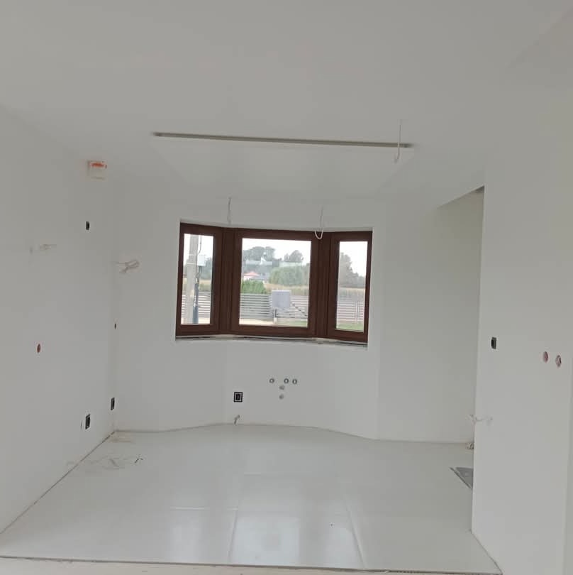 Wnętrze pokoju po gładziowaniu ścian i sufitu, zabezpieczona folią podłoga, okno z brązową ramą, widoczne puszki elektryczne.