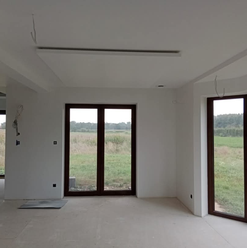 Wnętrze w stanie deweloperskim z białymi ścianami, brązowymi oknami z widokiem na łąkę, widoczne niedociągnięcia i brak wykończenia podłogi.