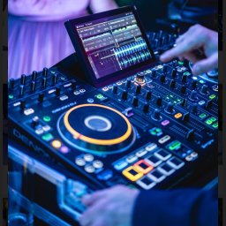 Adimat Mariusz Nowicki - DJ miksuje muzykę na profesjonalnym sprzęcie Denon DJ, na ekranie tabletu widoczne wykresy fal dźwiękowych, w tle fragment sukienki osoby.