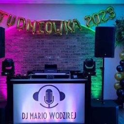 Adimat Mariusz Nowicki - Stanowisko DJ-a Mario Wodzireja na tle ceglanej ściany z balonami 'Studniówka 2023', oświetlone na różowo i niebiesko, z kolumnami głośnikowymi po bokach i dekoracjami z balonów w kolorach czarnym,...