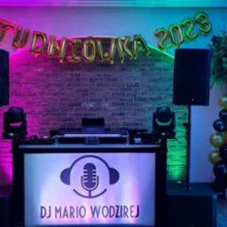 Stanowisko DJ-a Mario Wodzireja na tle ceglanej ściany z balonami 'Studniówka 2023', oświetlone na różowo i niebiesko, z kolumnami głośnikowymi po bokach i dekoracjami z balonów w kolorach czarnym,...