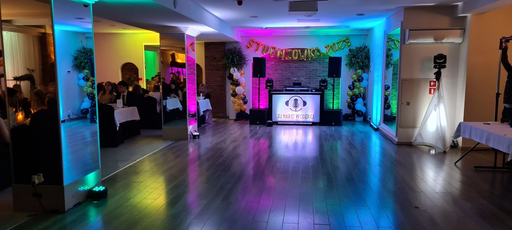 Sala studniówkowa w Bydgoszczy z dekoracjami balonowymi w kolorach czarnym, białym i złotym, oświetleniem LED w odcieniach różu, zieleni i niebieskiego oraz stanowiskiem DJ-a z napisem 'DJ Mario...