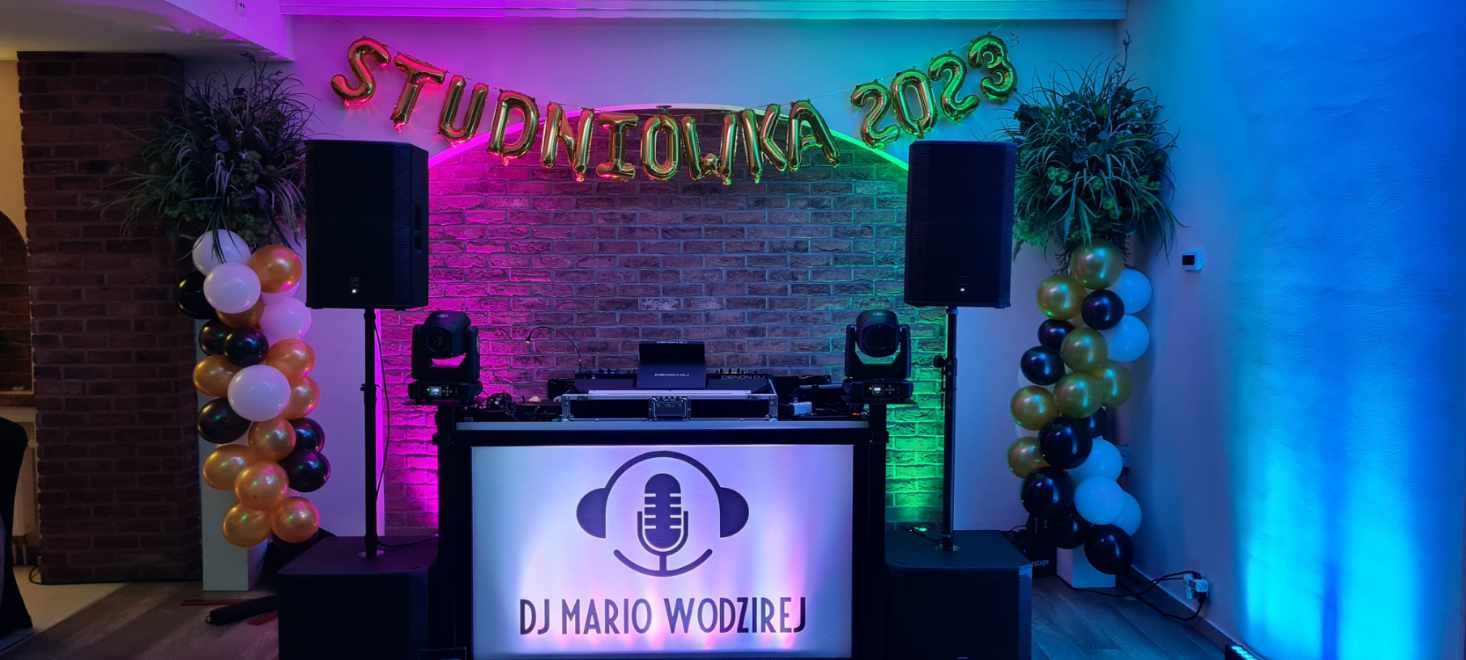 Stanowisko DJ-a Mario Wodzireja na tle ceglanej ściany z balonami 'Studniówka 2023', oświetlone na różowo i niebiesko, z kolumnami głośnikowymi po bokach i dekoracjami z balonów w kolorach czarnym,...