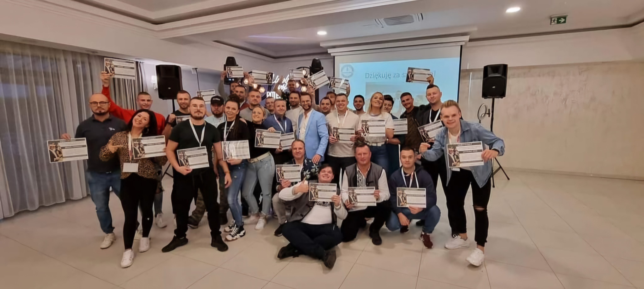 Grupa uśmiechniętych osób, trzymających certyfikaty na tle rzutnika z napisem 'Dziękuję za...', w jasnym pomieszczeniu z kolumną głośnikową