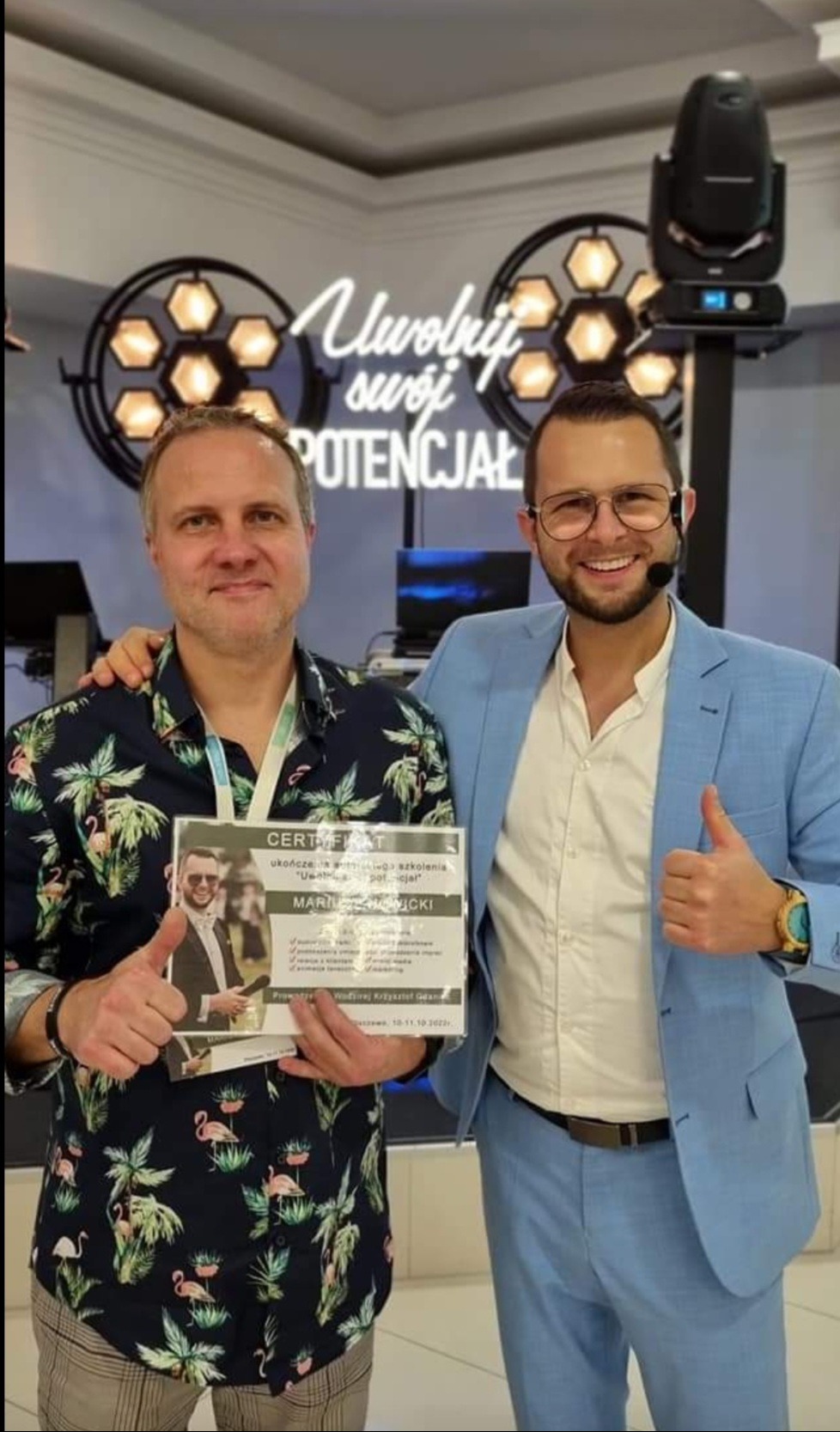 Dwóch mężczyzn pozuje z certyfikatem ukończenia szkolenia 'Uwolnij swój potencjał' na tle oświetlenia scenicznego i neonu z napisem 'Uwolnij swój potencjał'.