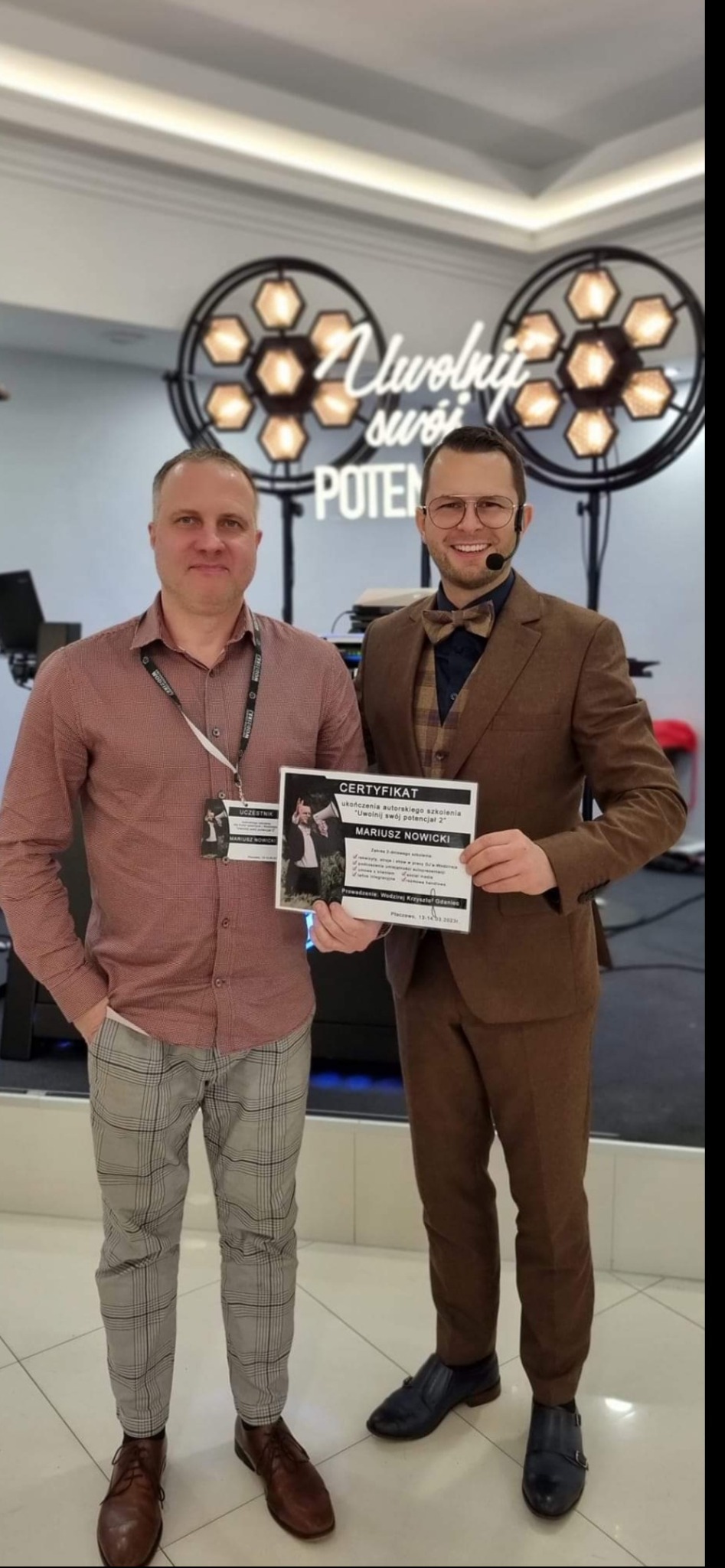 Dwaj mężczyźni prezentują certyfikat ukończenia szkolenia 'Uwolnij swój potencjał 2', na tle dekoracji świetlnej z napisem 'Uwolnij swój potencjał'.