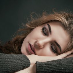 Foto Kubiczek - Portret śpiącej kobiety z delikatnym makijażem oczu, opierającej głowę na skrzyżowanych ramionach w szarym swetrze na ciemnym tle.
