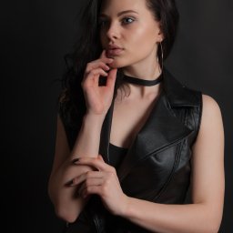 Foto Kubiczek - Portret młodej kobiety w czarnej, skórzanej kamizelce, z dłonią pod brodą, patrzącej w lewo, na ciemnym tle.
