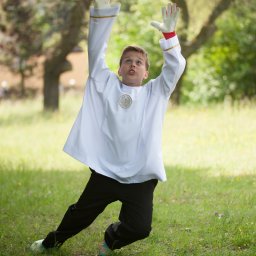 Foto Kubiczek - Chłopiec w albie komunijnej łapie piłkę na łące, w tle drzewa.