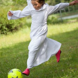 Foto Kubiczek - Dziewczynka w białej albie i różowych butach kopie żółto-czarną piłkę na zielonej trawie, rozłożone ramiona dla równowagi.
