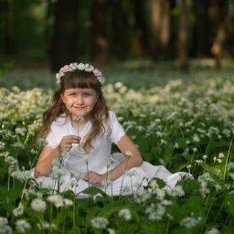 Foto Kubiczek - Dziewczynka w białej sukience i wianku siedzi pośród kwitnących białych kwiatów w lesie, trzymając pojedynczy kwiat.